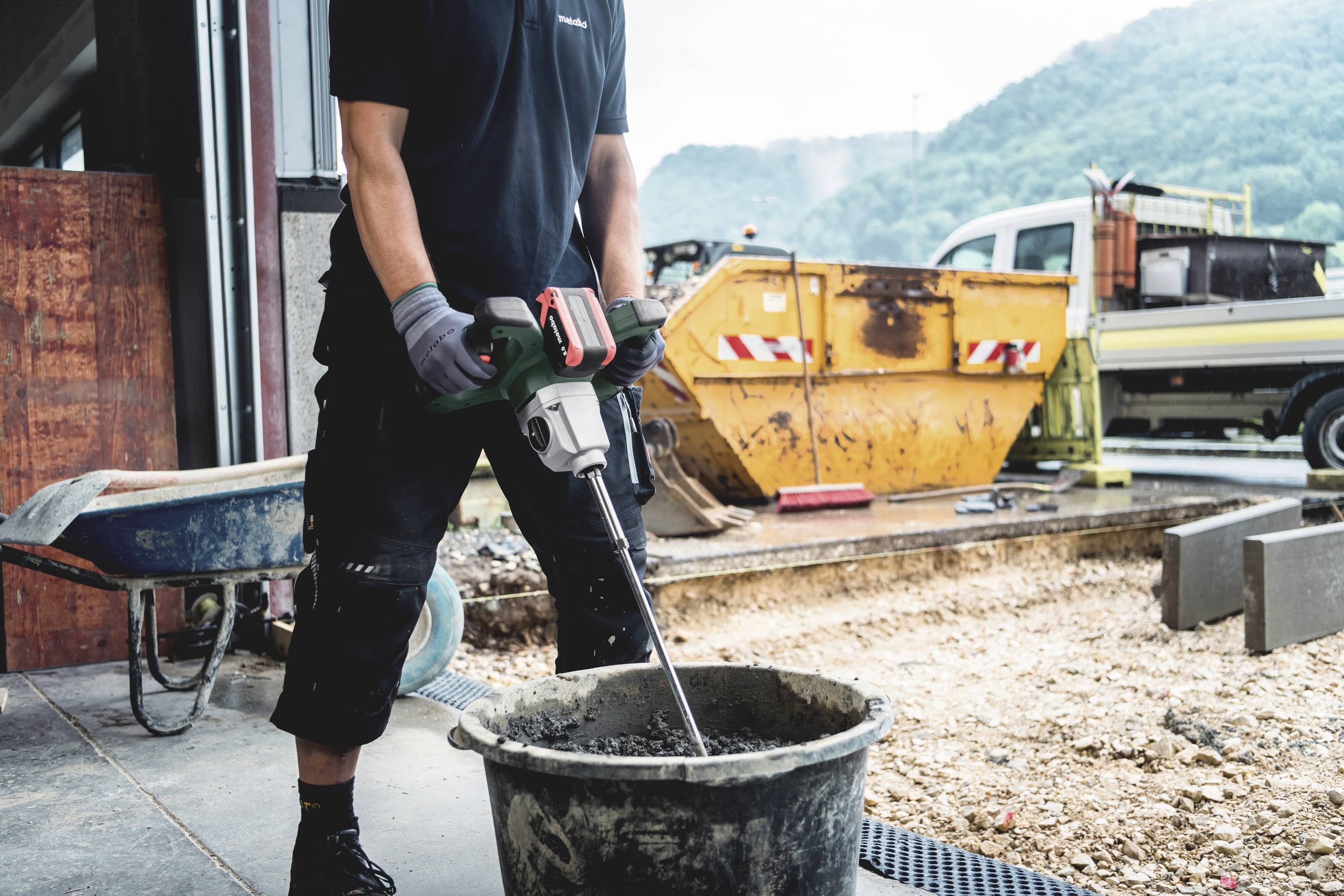 Arbeiter mischt Beton mit einem Rührwerk auf einer Baustelle, im Hintergrund Container und Lkw.