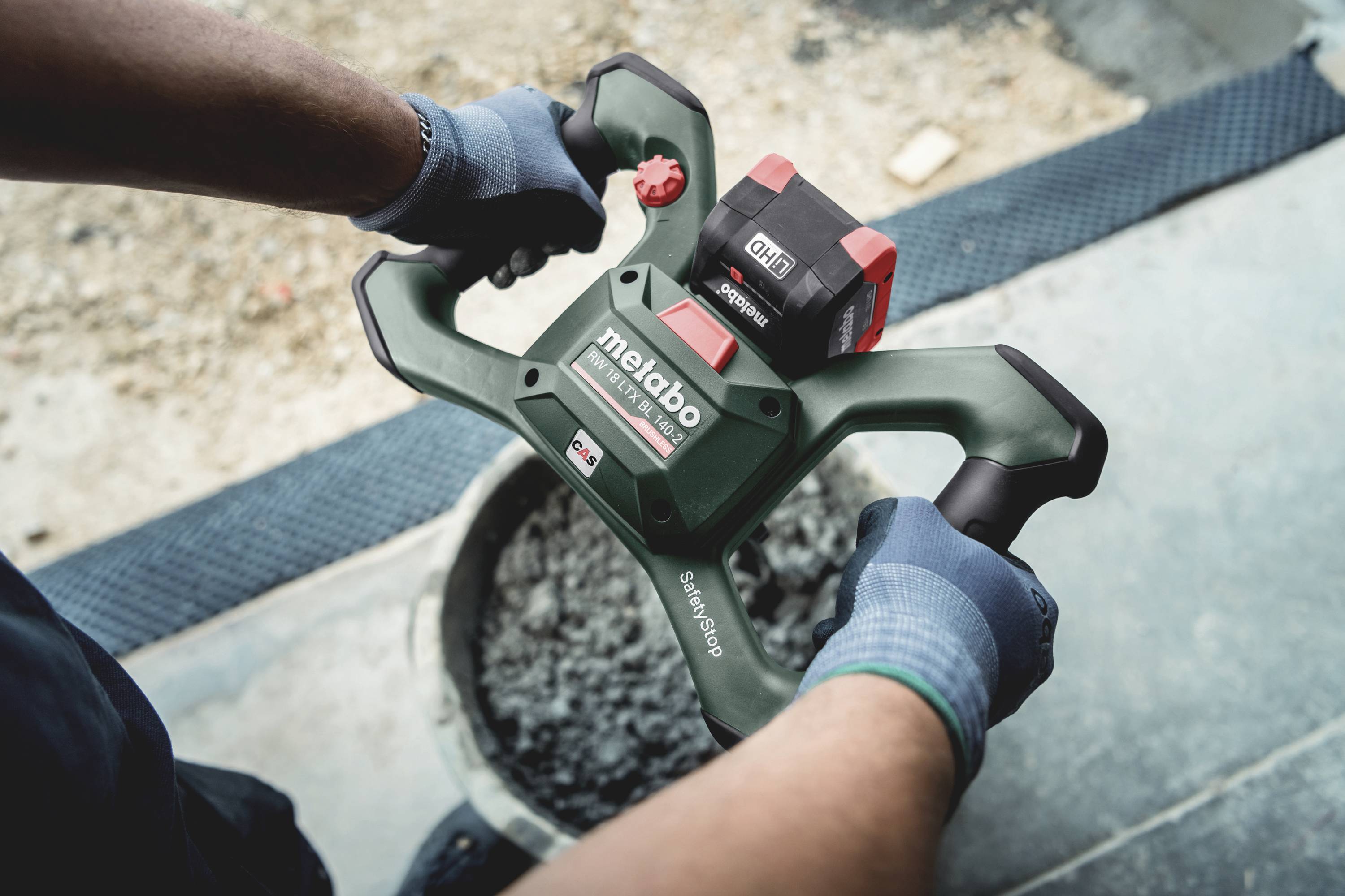 Person hält einen elektrischen Betonmischer der Marke 'Metabo' über einen Eimer mit Betonmischung auf einer Baustelle.