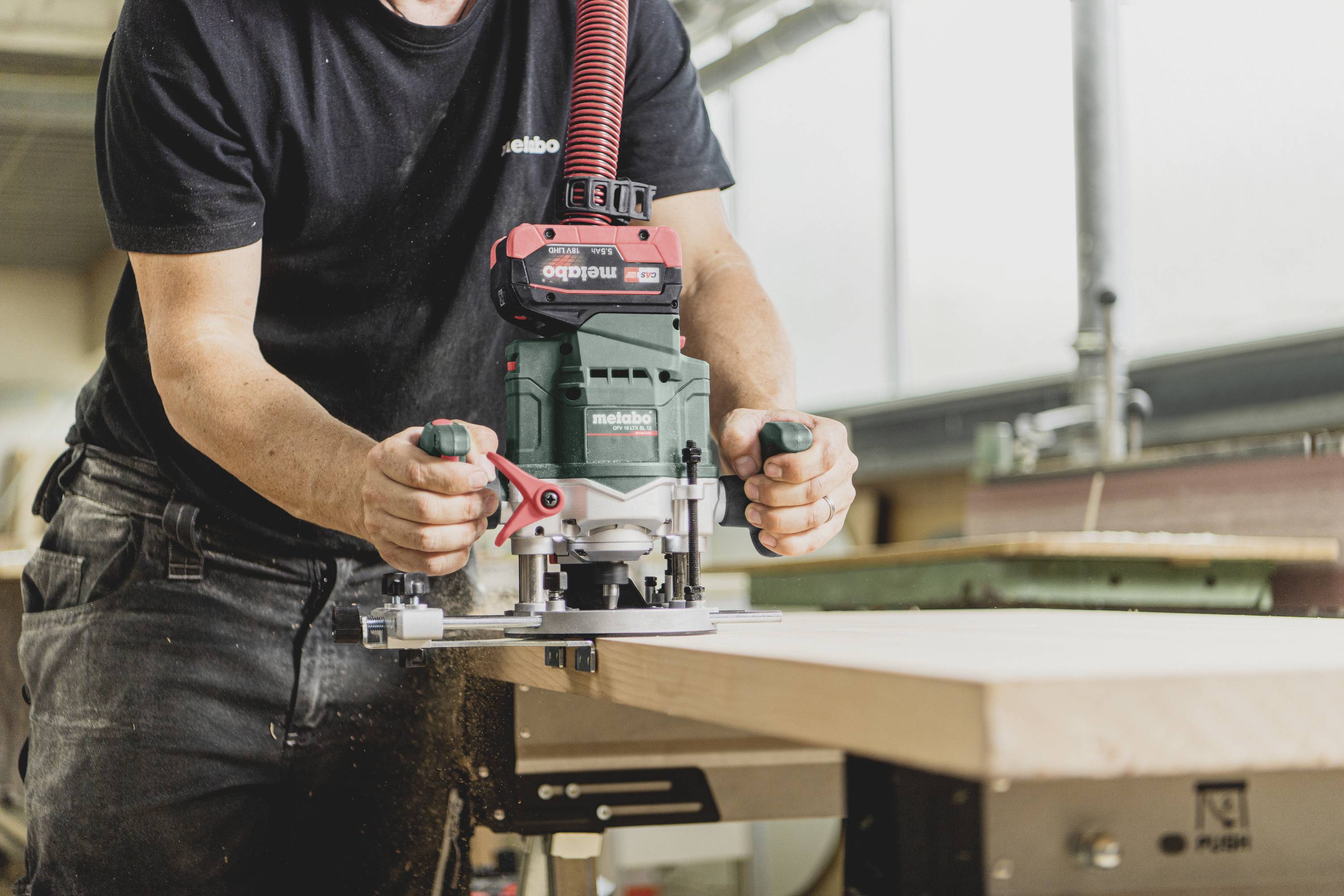 Eine Person arbeitet mit einer Fräse, um Holz in einer Werkstatt zu bearbeiten. Die Umgebung ist hell und es herrscht handwerkliche Aktivität.