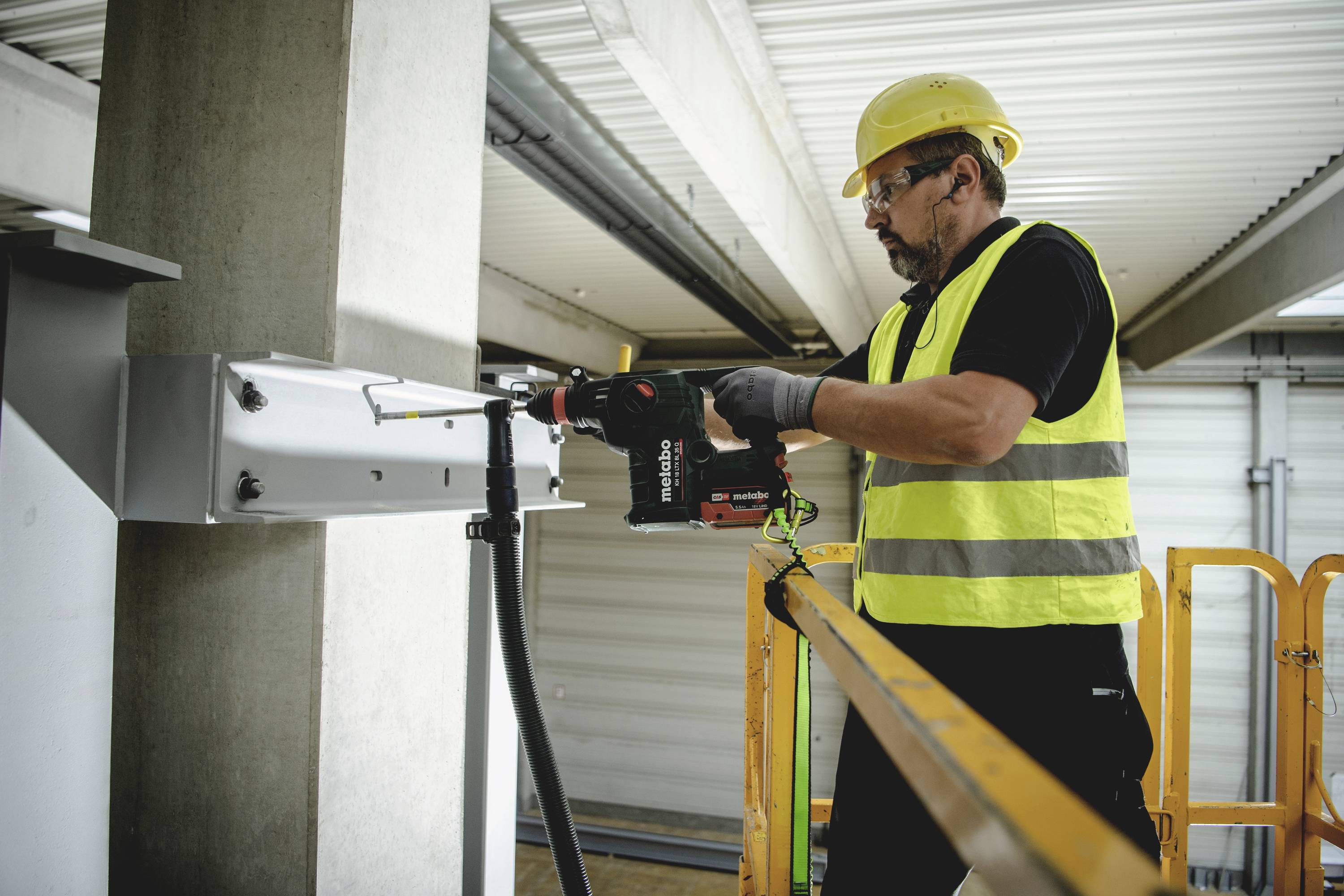 Ein Bauarbeiter mit gelber Weste und Helm verwendet eine elektrische Bohrmaschine, um an einer Metallkonstruktion zu arbeiten.