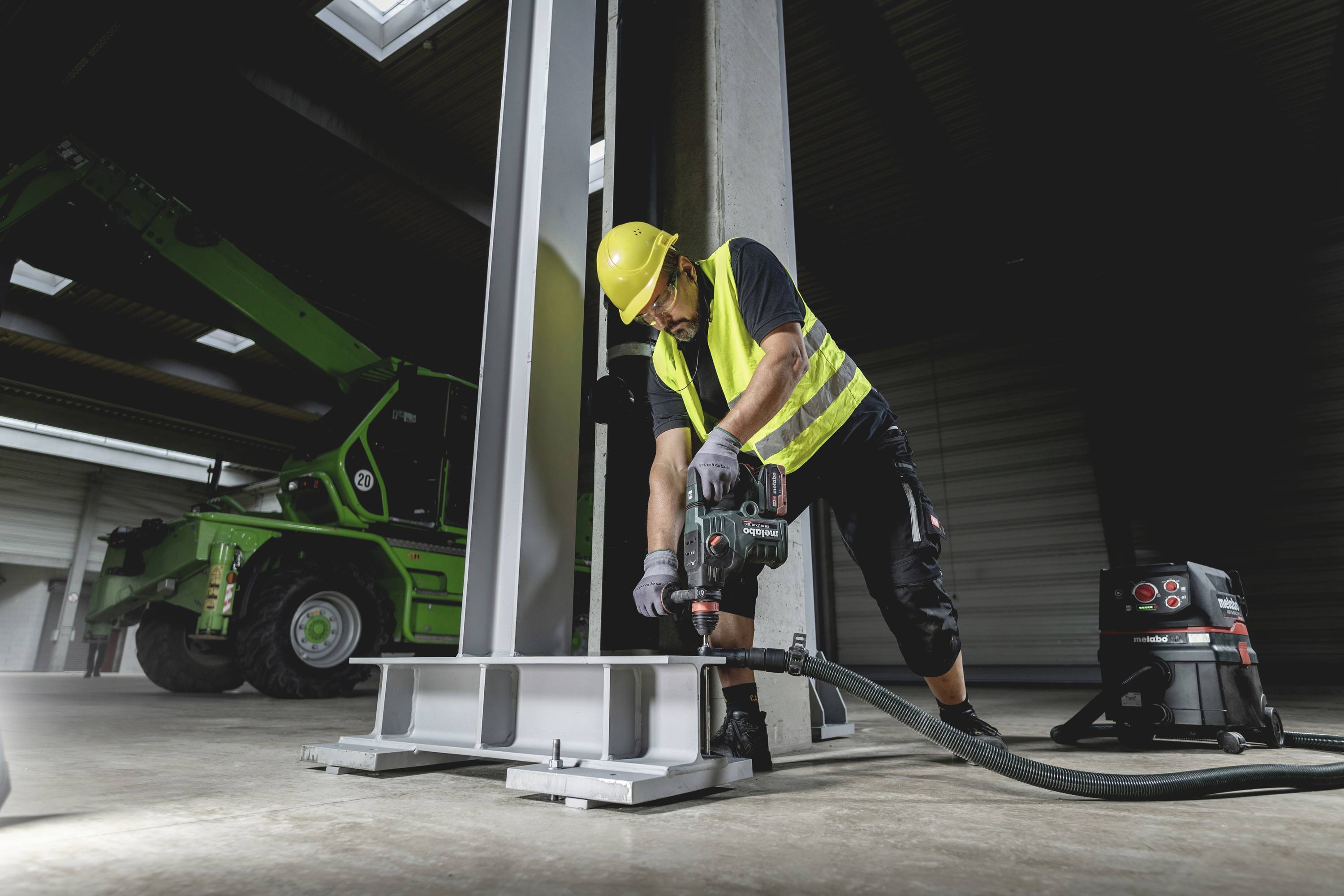 Ein Bauarbeiter mit gelber Weste und Helm bohrt Metall in einer Halle. Ein grüner Kran im Hintergrund.