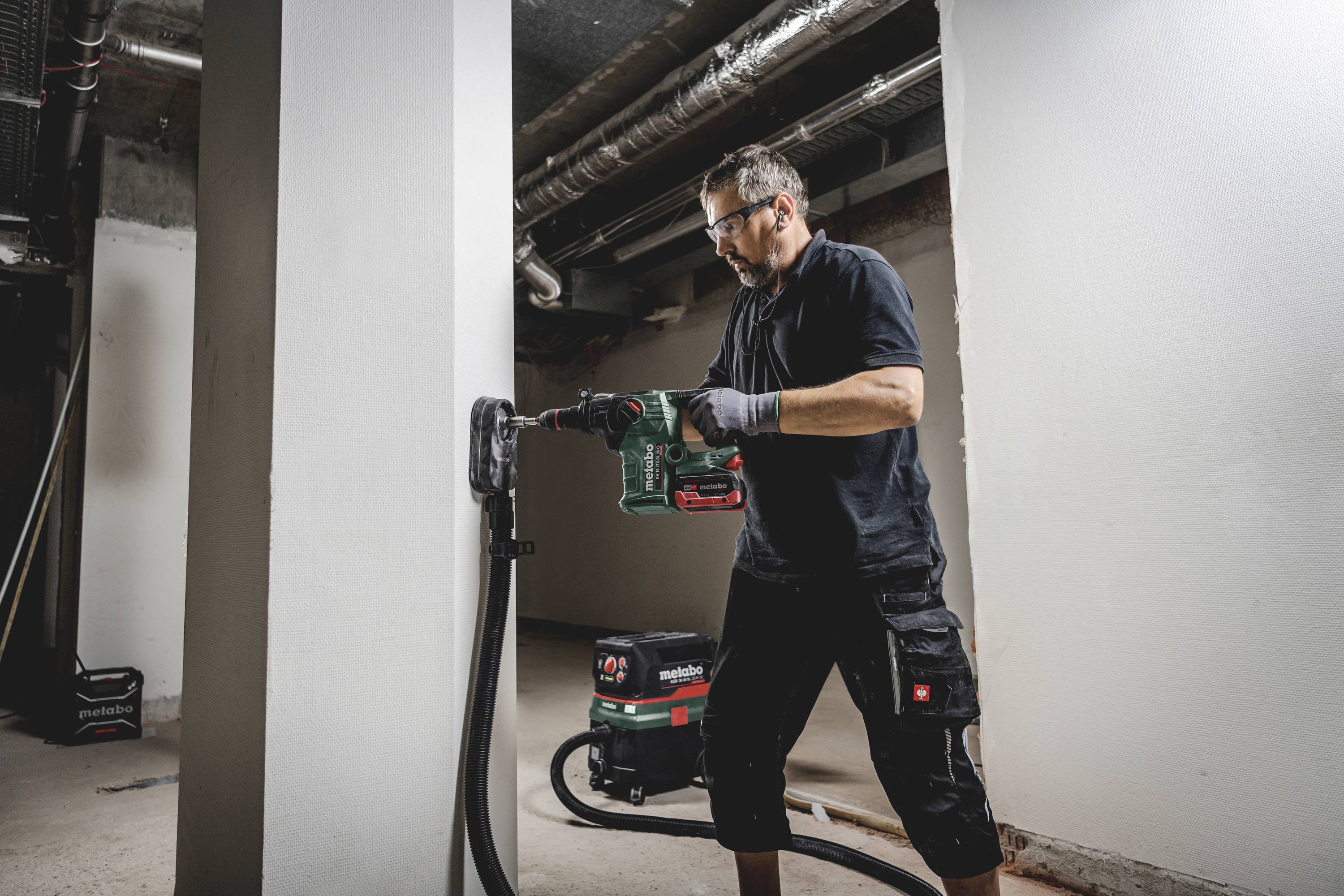 Ein Mann in Arbeitskleidung bohrt mit einer grünen Bohrmaschine in eine Wand in einem unfertigen Raum. Ein Staubsauger steht neben ihm.