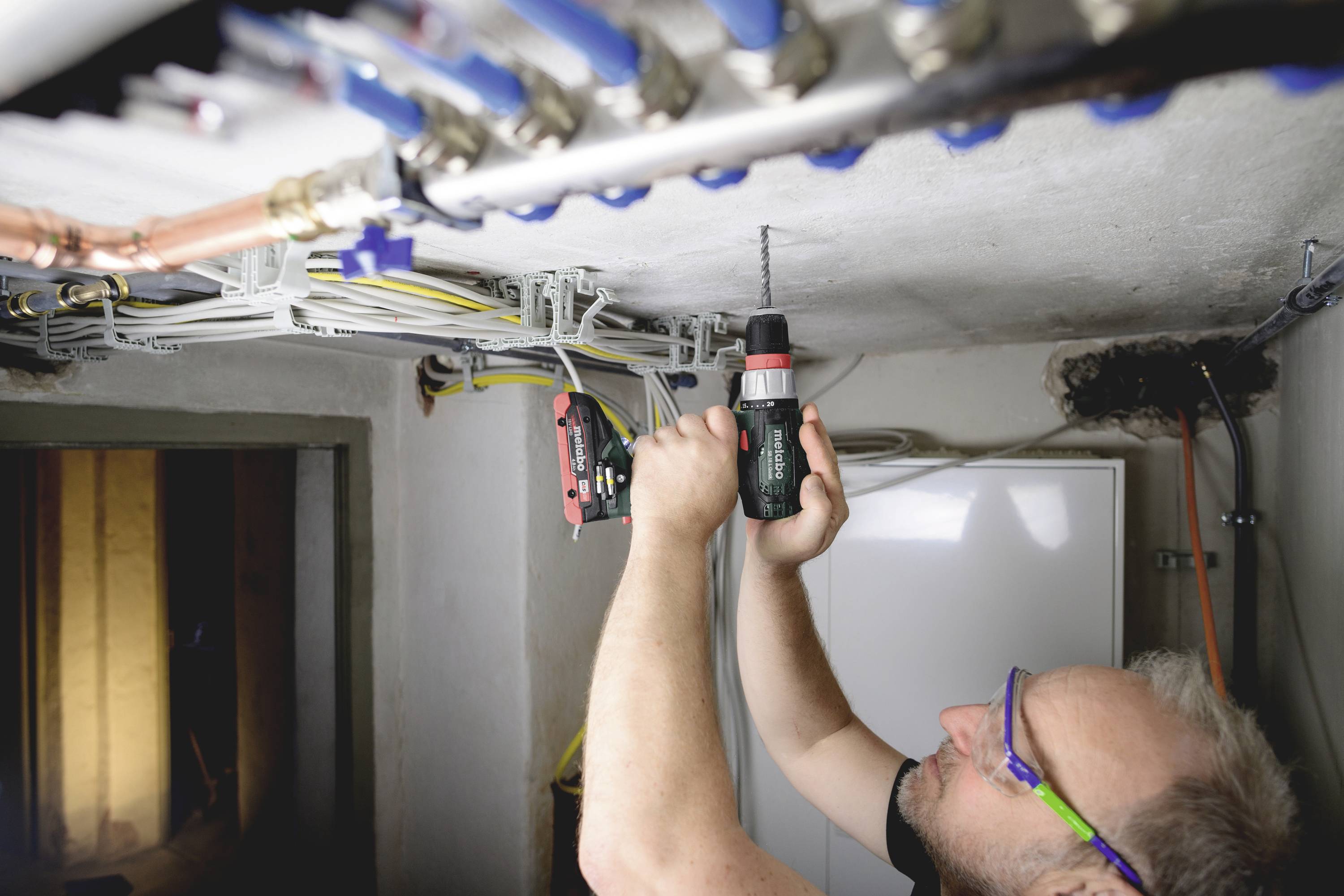Ein Handwerker bohrt ein Loch in die Zimmerdecke. Über ihm verlaufen zahlreiche farbige Rohre und Kabel. Der Raum wirkt industriell.