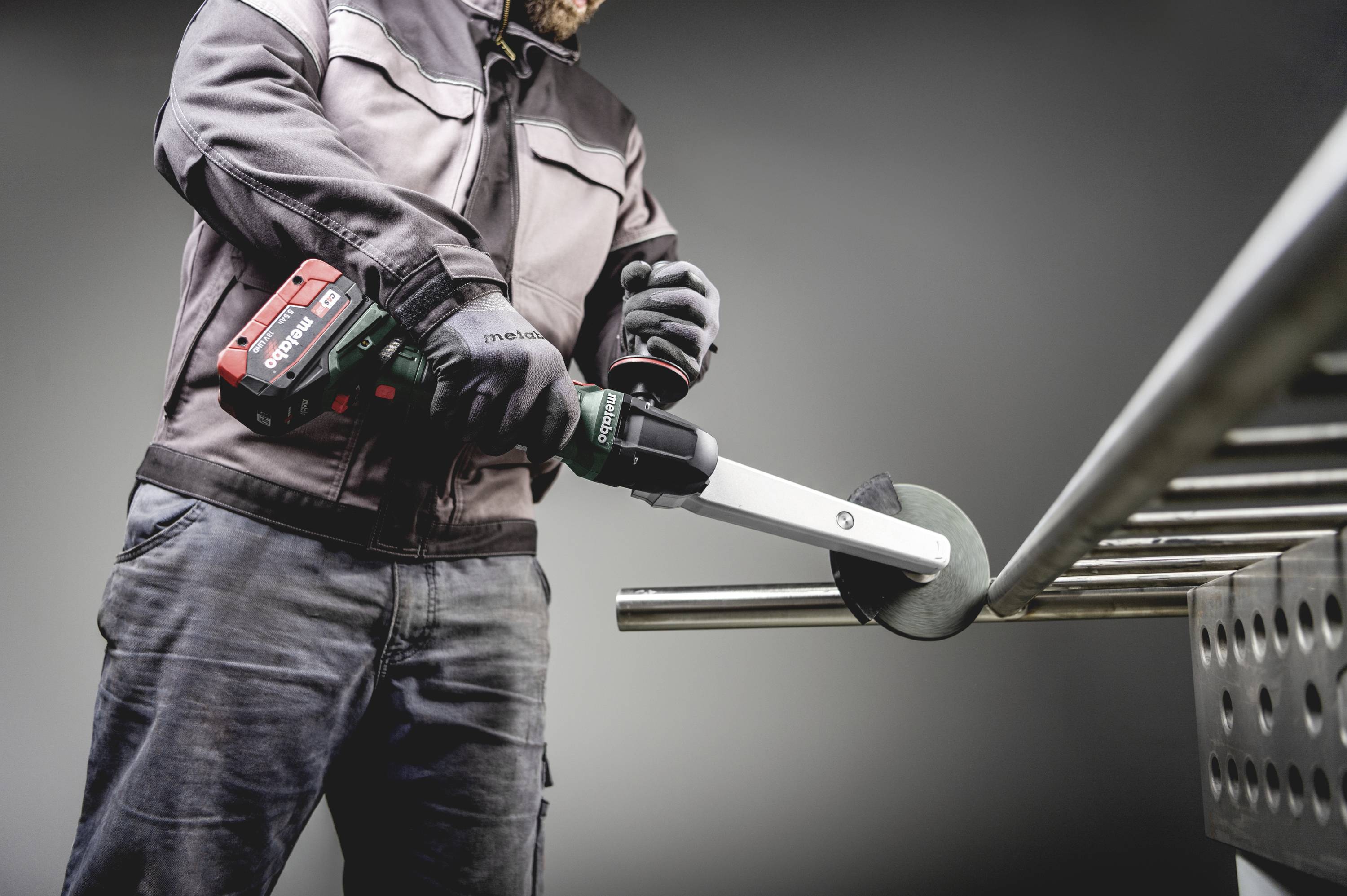 Ein Arbeiter schleift ein Metallrohr mit einem elektrischen Winkelschleifer. Er trägt Schutzkleidung und Handschuhe.