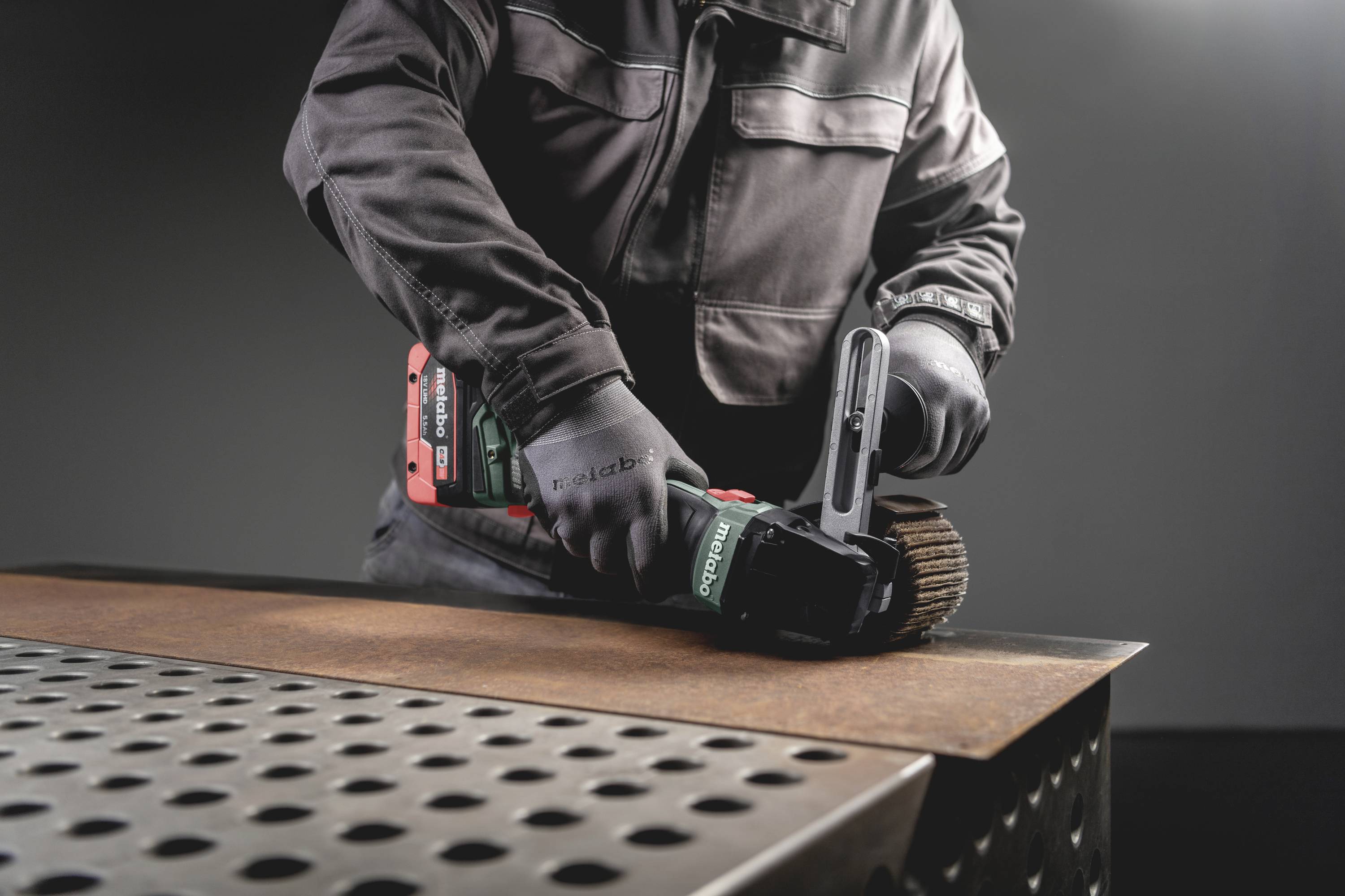Eine Person schleift mit einem elektrischen Schleifgerät eine Metallplatte auf einem Tisch mit Lochmuster. Sie trägt Schutzhandschuhe.
