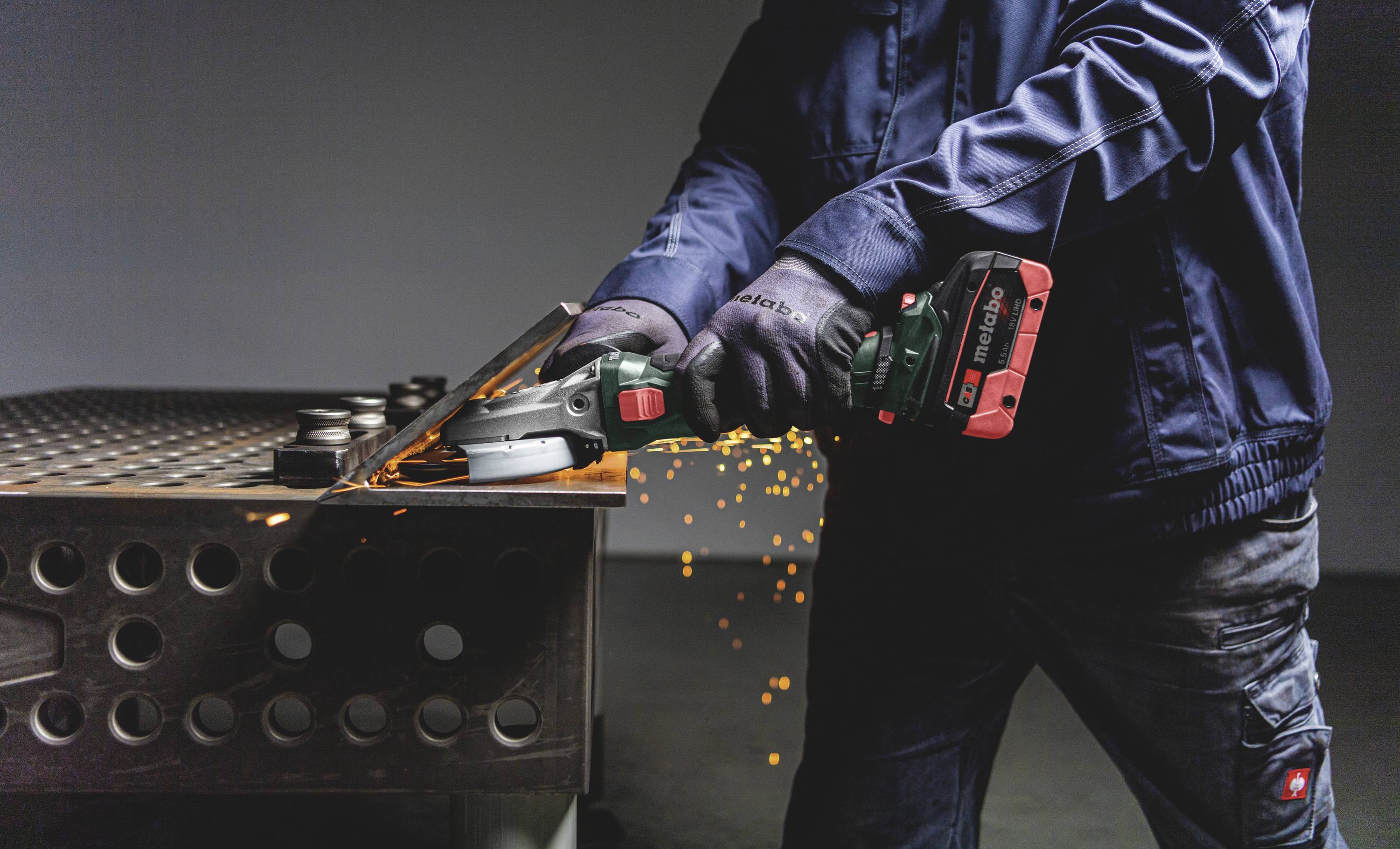 Eine Person schneidet Metall mit einem Winkelschleifer, Funken fliegen. Fokus auf das Werkzeug und die Arbeitssituation.