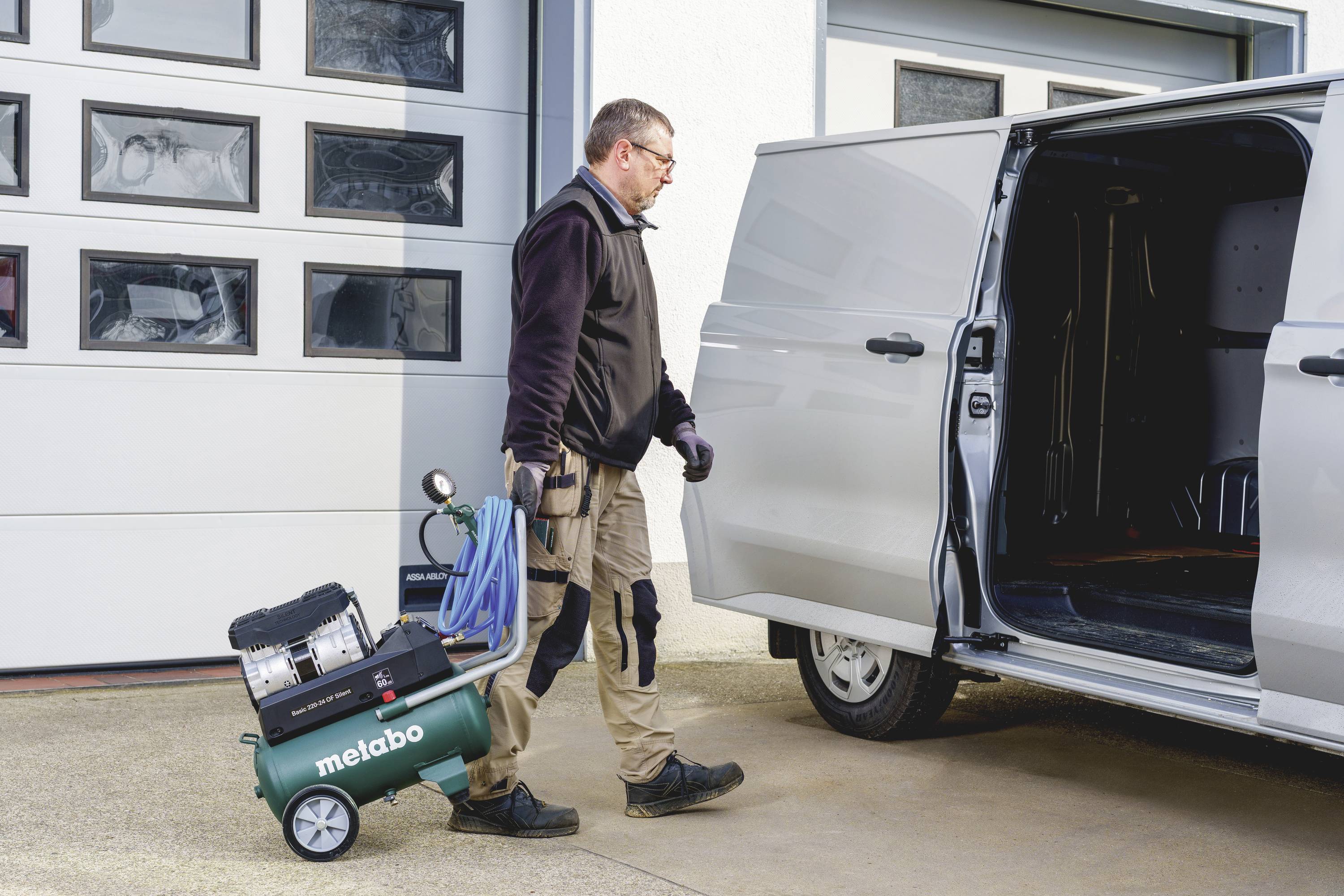Ein Mann lädt einen grünen Metabo-Kompressor in einen Lieferwagen vor einer Garage. Er trägt Arbeitskleidung und Handschuhe.