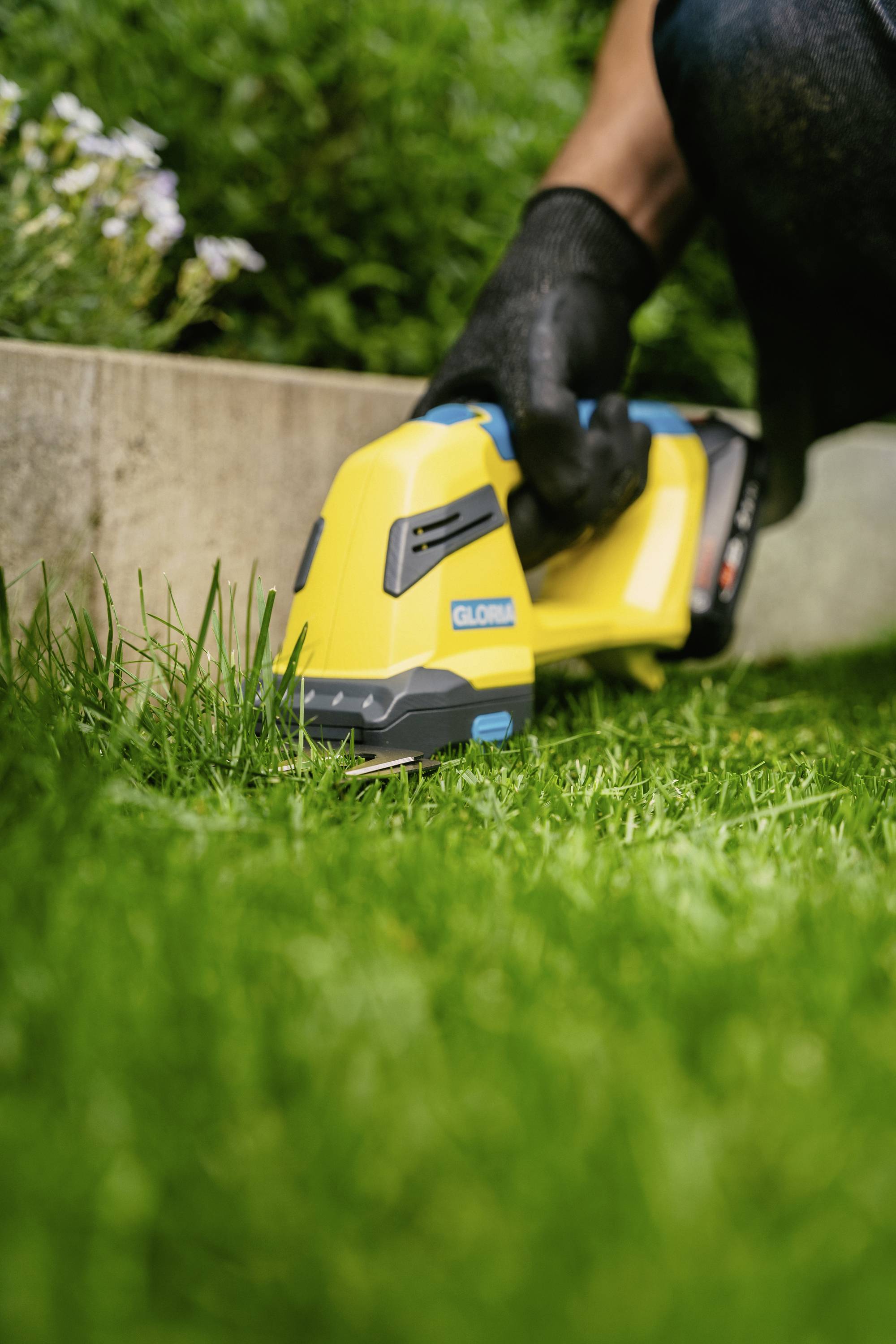 Person schneidet Gras mit einem gelben elektrischen Rasentrimmer in einem Garten. Fokus auf Gerät und gemähten Rasen.