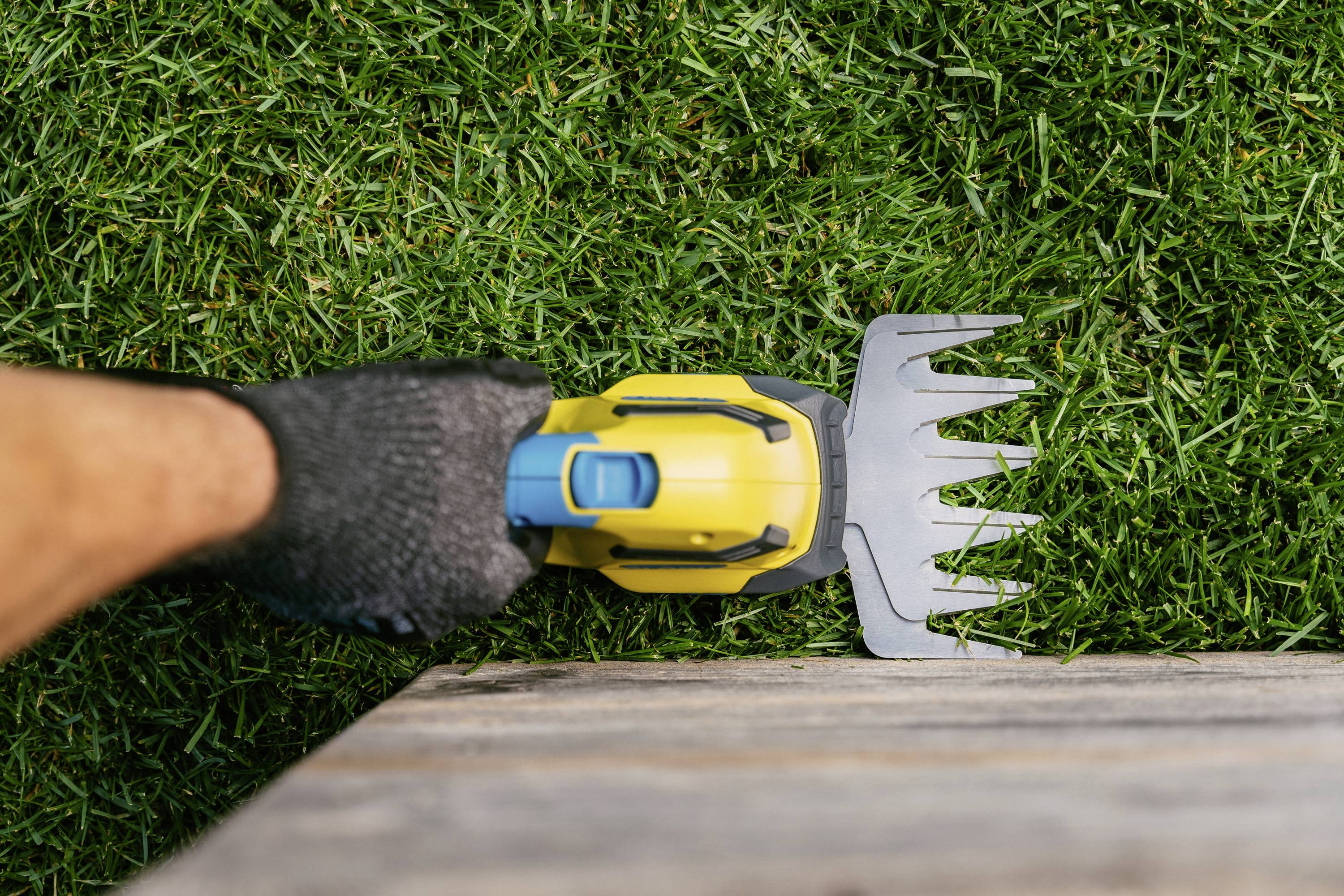Ein Handschneider mit gelbem Griff wird verwendet, um Rasen an einer Holzkante präzise zu trimmen. Eine behandschuhte Hand hält das Werkzeug.