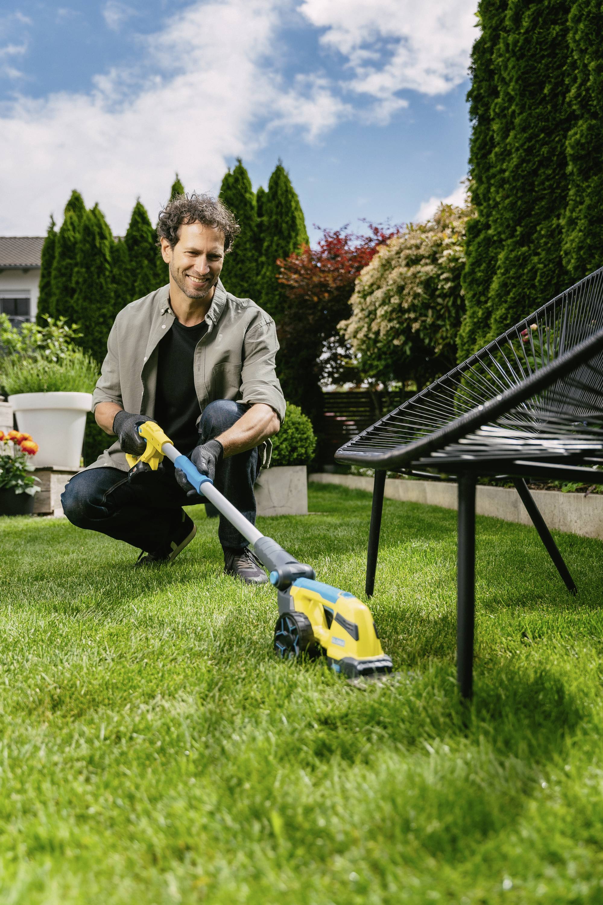 Ein Mann mäht den Rasen mit einem elektrischen Rasenmäher in einem gepflegten Garten mit Büschen und Pflanzen im Hintergrund.