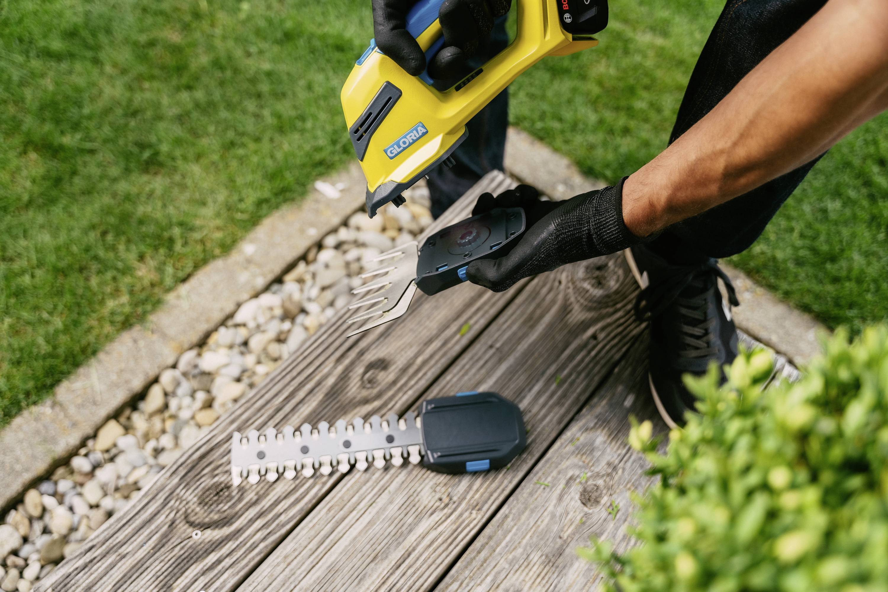 Eine Person wechselt die Klingen eines gelben Gartentrimmerschneiders, der auf der Terrasse neben einem kleinen Gebüsch liegt.