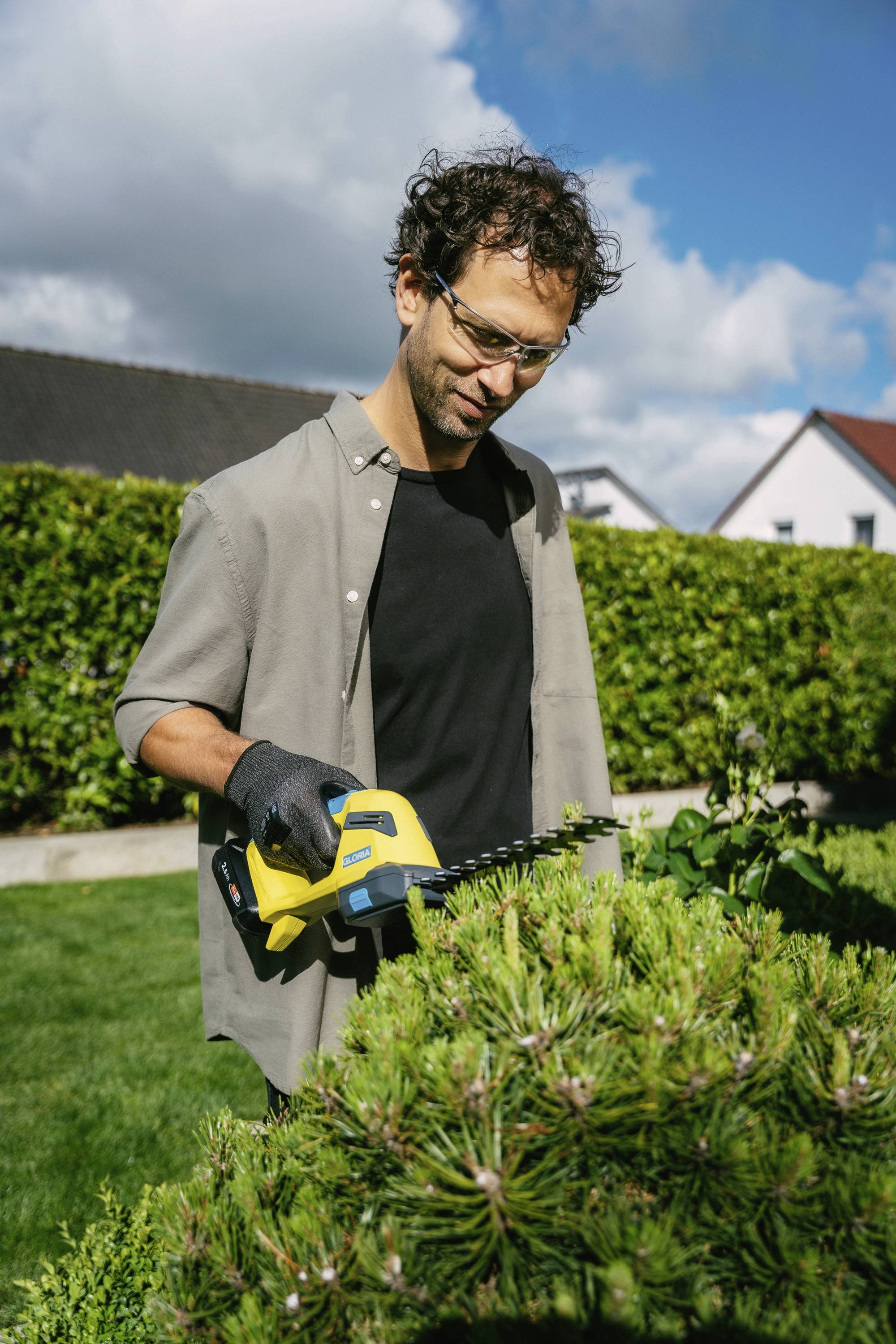 Ein Mann trägt eine Schutzbrille und schneidet mit einer elektrischen Heckenschere eine Hecke im Garten.