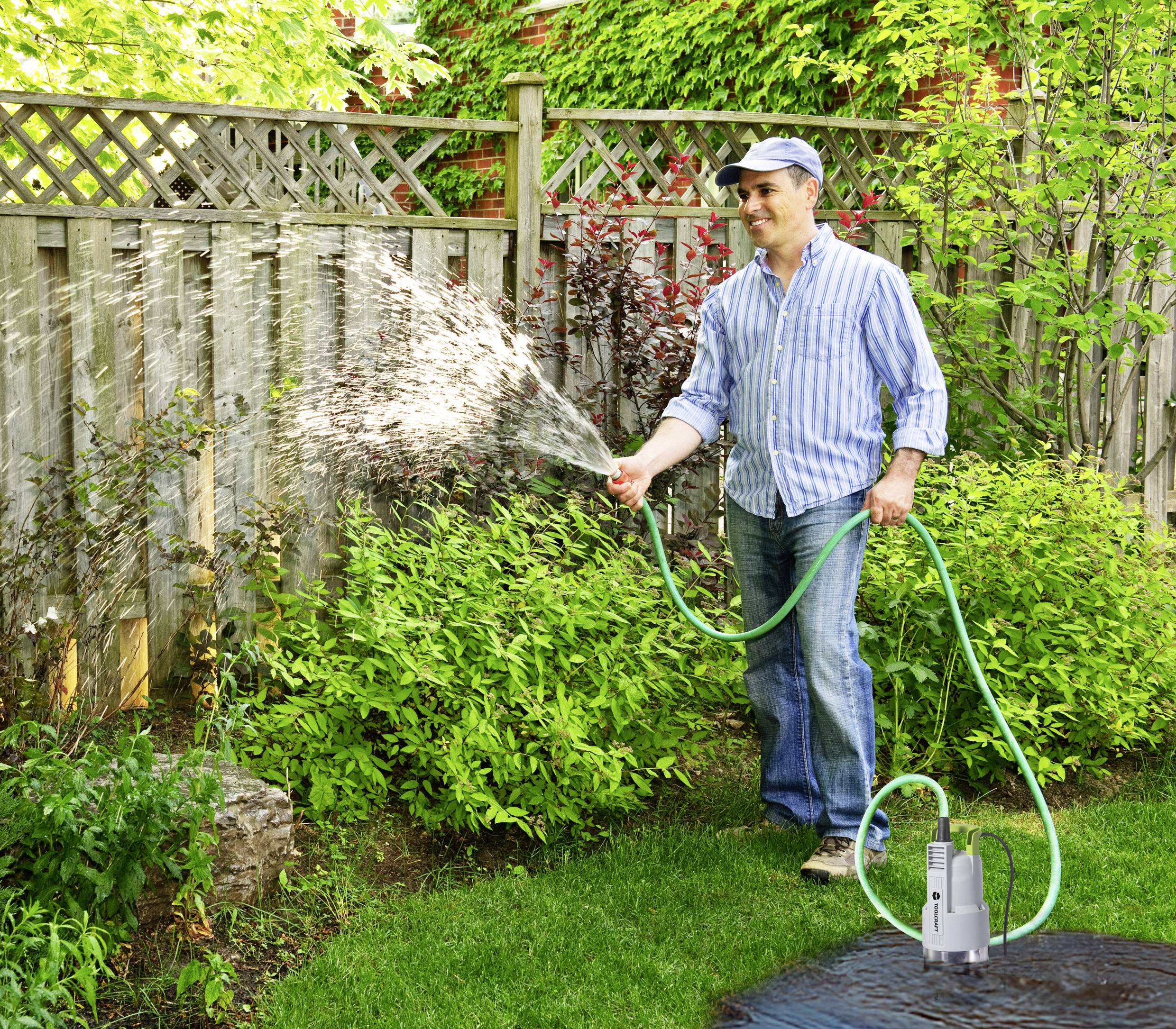 Ein Mann bewässert den Garten mit einem grünen Schlauch. Er steht auf einer Rasenfläche, umgeben von Pflanzen und einem Holzzaun.
