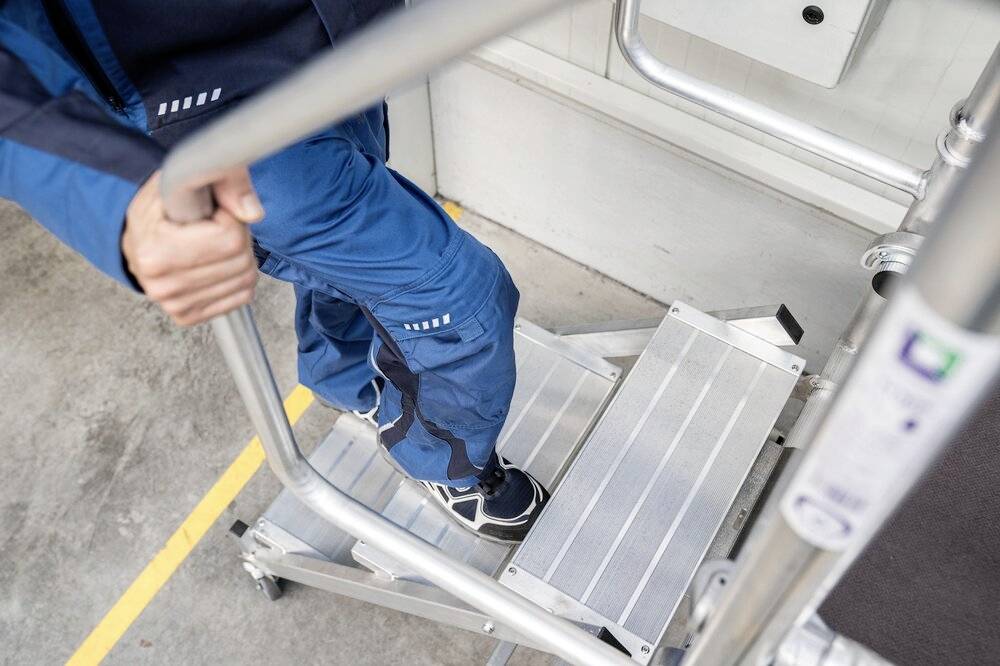 Eine Person in blauer Arbeitskleidung steigt eine mobile Aluminiumtreppe hinauf. Die Treppe hat rutschfeste Stufen.
