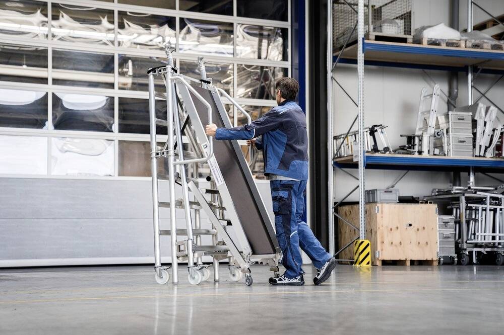 Eine Person schiebt eine fahrbare Aluminiumleiter in einer Lagerhalle. Im Hintergrund sind Regale mit Metallteilen zu sehen.