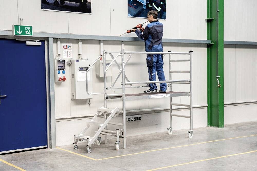 Eine Person in Arbeitskleidung steht auf einem mobilen Gerüst in einer Industriehalle und arbeitet mit einem Werkzeug an der Wand.