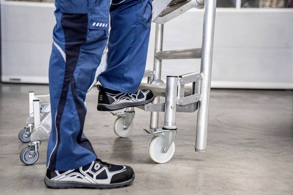 Eine Person in blauer Arbeitshose steigt auf eine Leiter mit Rollen. Der Boden ist grau und glat, was auf eine Werkstattumgebung hindeutet.