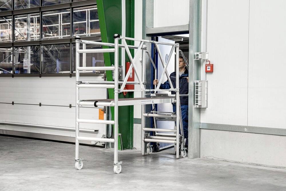 Eine Person schiebt eine faltbare Aluminium-Plattform auf Rollen durch eine Tür in einer Industriehalle. Die Plattform ist teilweise zusammengeklappt.