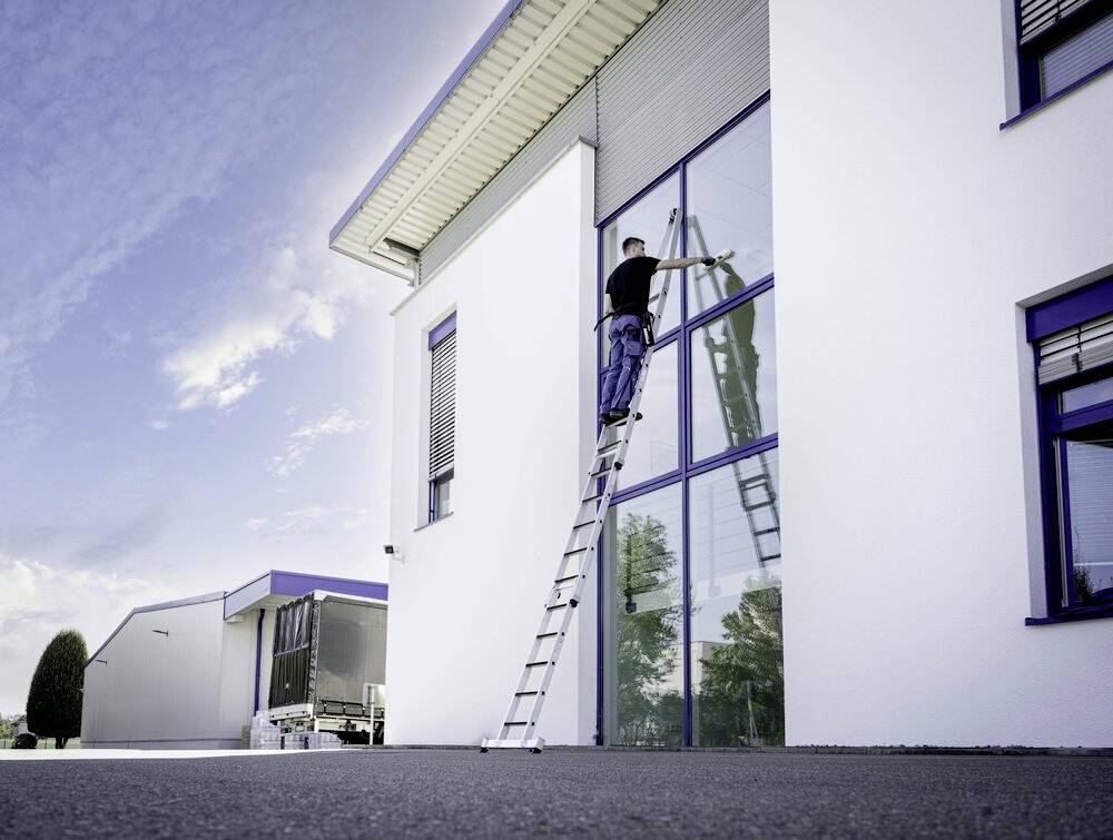 Mann steht auf Leiter und putzt große Fenster eines modernen Gebäudes. Blauer Himmel im Hintergrund, andere Gebäude sichtbar.