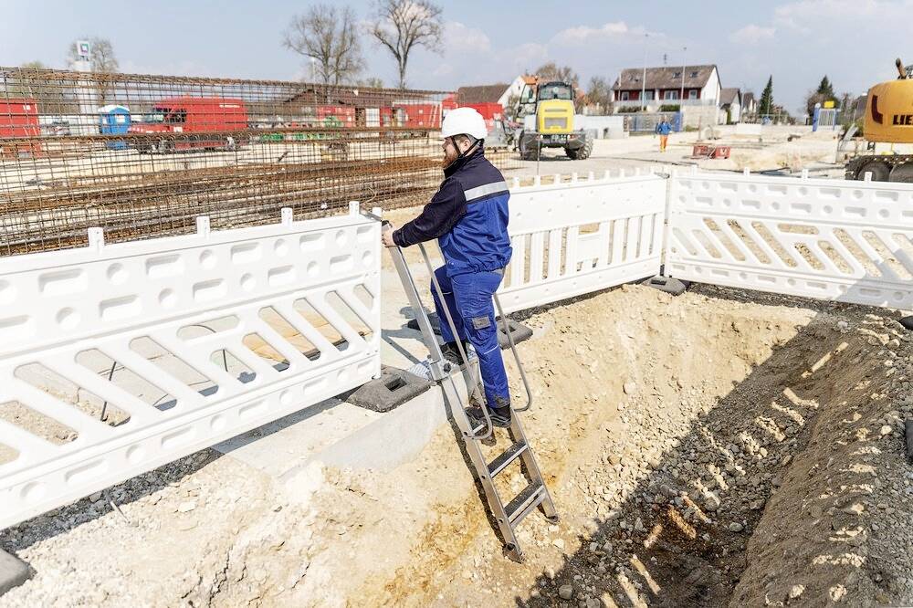 Eine Person in Baukleidung steigt mit einem Helm eine Leiter hinunter in eine Baugrube. Im Hintergrund sind Bauzäune und Gebäude sichtbar.