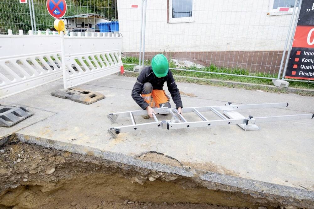 Eine Person mit grünem Schutzhelm baut eine Leiter bei einer Baustelle neben einem Graben auf. Im Hintergrund ist ein Bauzaun zu sehen.