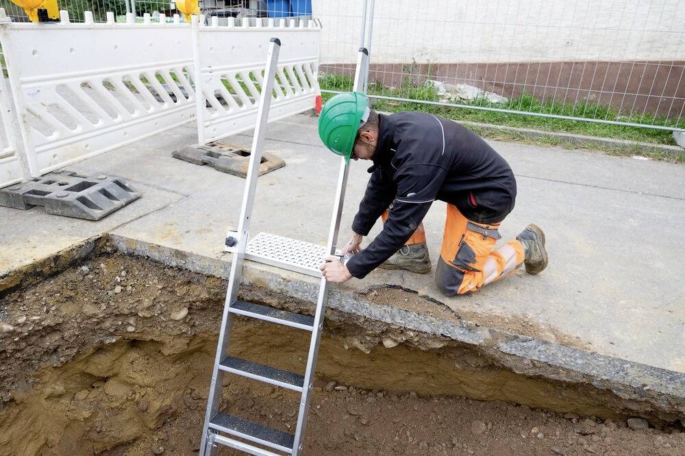 Arbeiter mit Schutzhelm kniet an einer Baustelle und sichert eine Leiter am Rand eines Grabens.