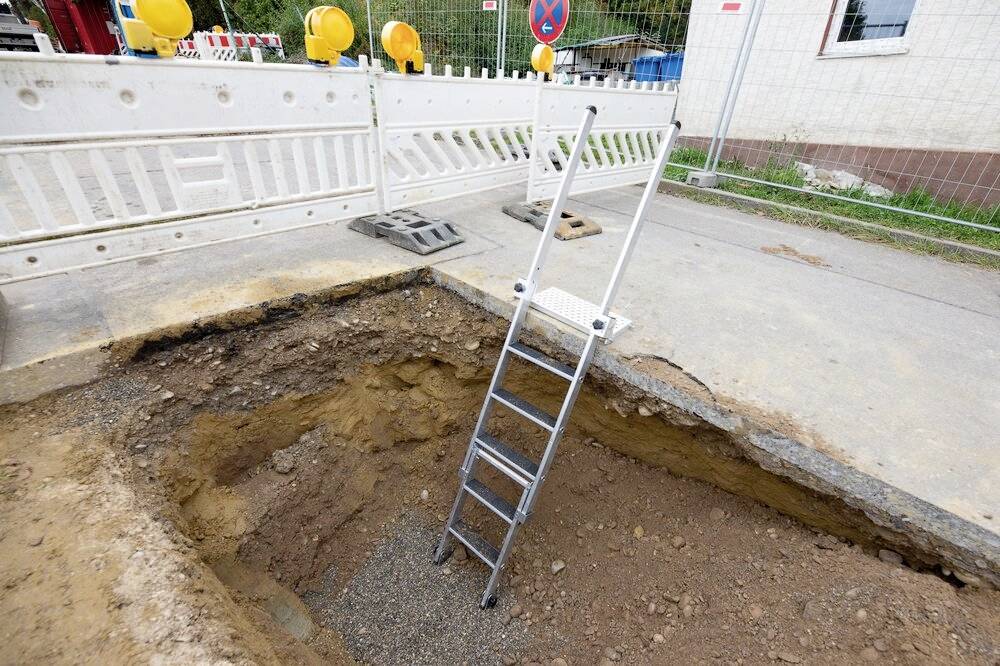 Baugrube auf einer Straße mit einer Leiter, die hineinführt. Absperrungen mit Warnschildern im Hintergrund, die Baustelle absichern.