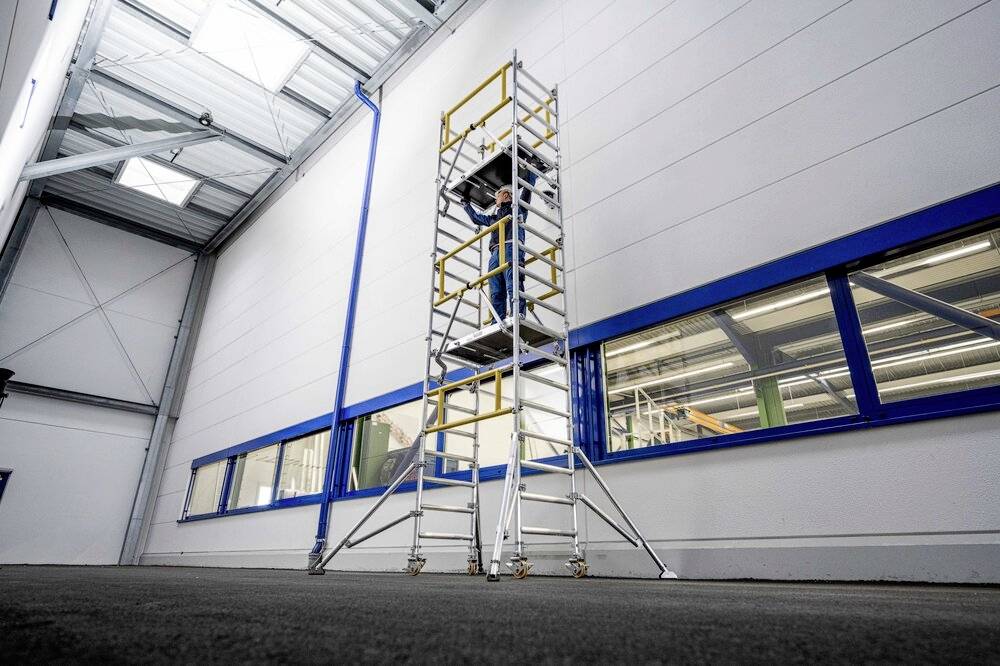 Eine Person steht auf einem hohen Gerüst in einer großen Halle. Das Gerüst ist aus Metall mit gelben Sicherheitselementen montiert.