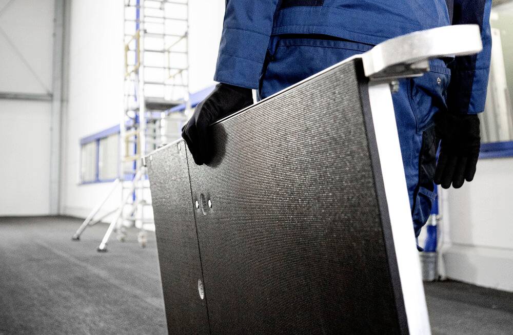 Ein Arbeiter in blauer Arbeitskleidung trägt eine große rechteckige Platte durch einen industriellen Raum mit Gerüst im Hintergrund.