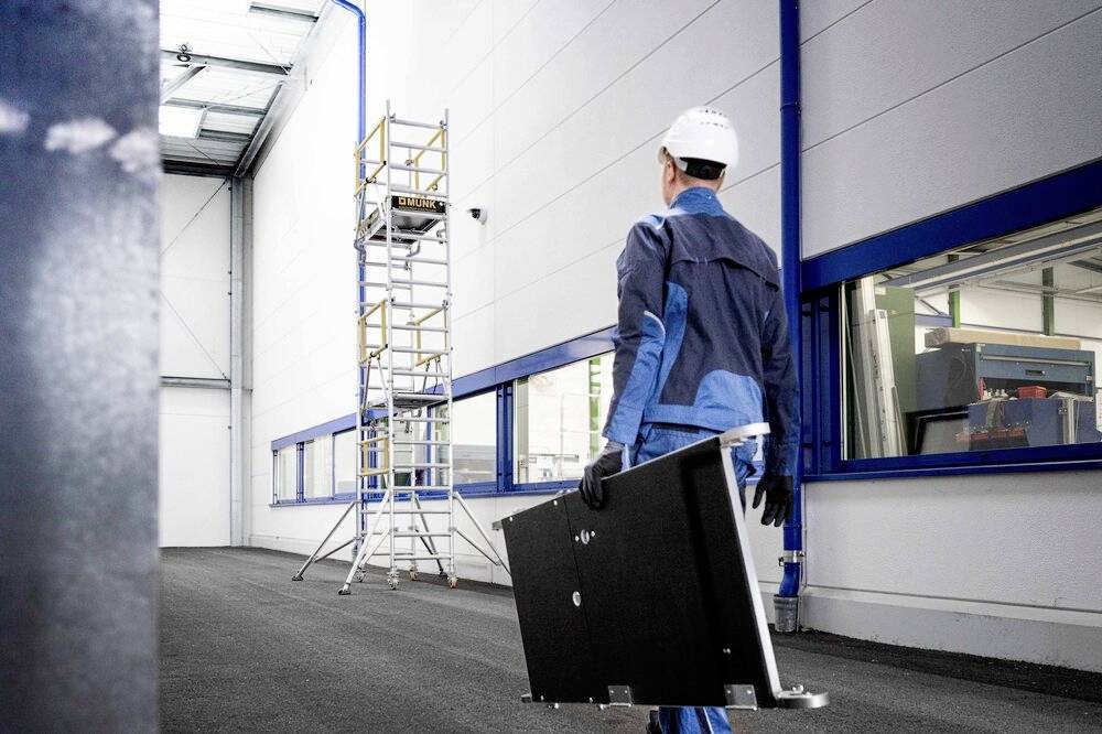 Ein Arbeiter in blauer Schutzkleidung und Helm trägt ein schwarzes Paneel in einer hellen Werkshalle, im Hintergrund ein Gerüst.