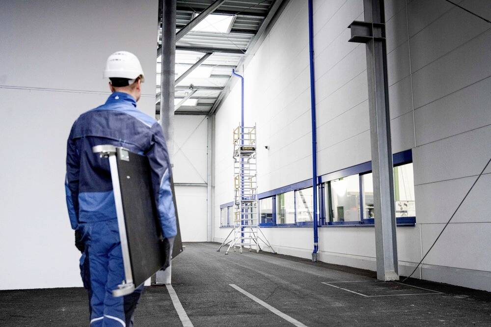 Ein Arbeiter mit Schutzhelm trägt ein Solarpanel entlang eines Außenkorridors einer Industrieanlage, im Hintergrund steht ein Gerüst.