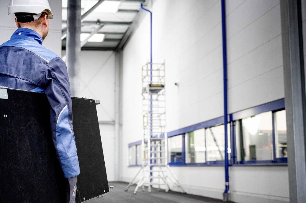 Ein Arbeiter in Schutzkleidung trägt eine Metallplatte in einer Fabrikhalle. Im Hintergrund ist ein Gerüst an der Wand zu sehen.