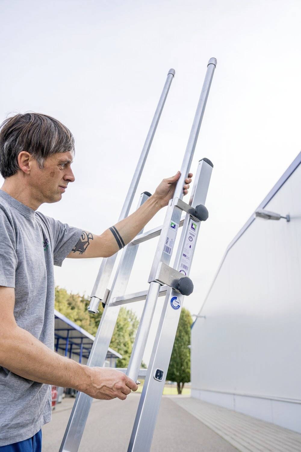 Ein Mann stellt eine lange Leiter vor einem Gebäude auf. Er trägt ein graues T-Shirt und konzentriert sich auf die Leiter.