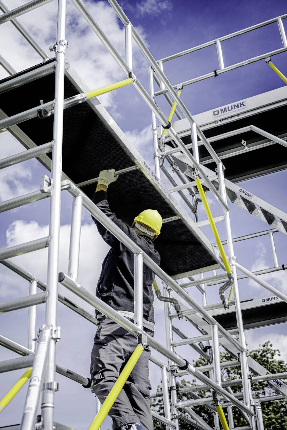 Arbeiter mit Helm hebt Metallplatte auf ein Gerüst unter blauem Himmel, Sicherheitskonstruktion deutlich sichtbar.