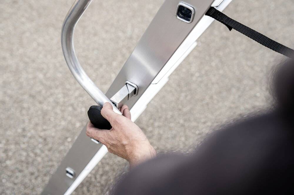 Eine Person hält die Seite einer Leiter und zieht an einem Hebel. Die Leiter ist auf einem grobkörnigen, grauen Hintergrund positioniert.