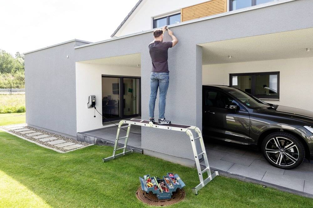 Ein Mann steht auf einer Leiter und arbeitet am Dach eines modernen Hauses. Unten ist ein offenes Werkzeugkasten und eine geparkte Limousine sichtbar.