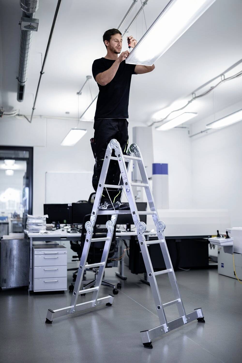 Eine Person steht auf einer Leiter in einem modernen Büro und wechselt eine Leuchtstofflampe an der Decke.