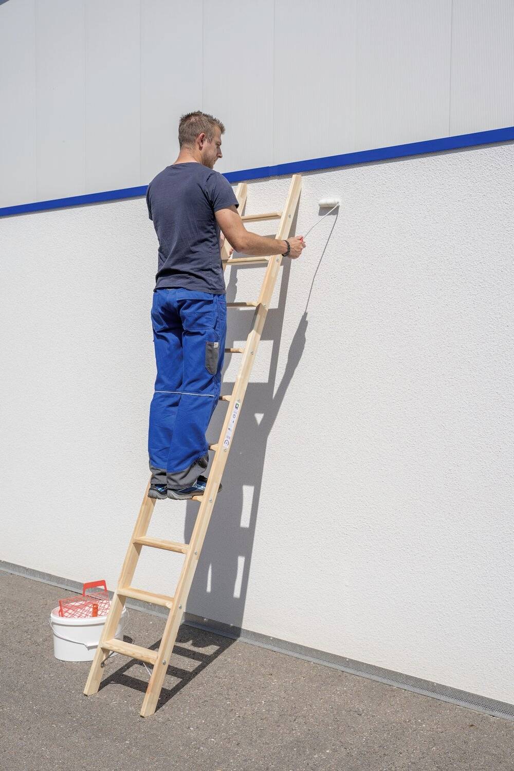 Ein Mann in Arbeitskleidung steht auf einer Leiter und streicht eine weiße Wand blau. Unten steht ein Eimer mit Farbe.