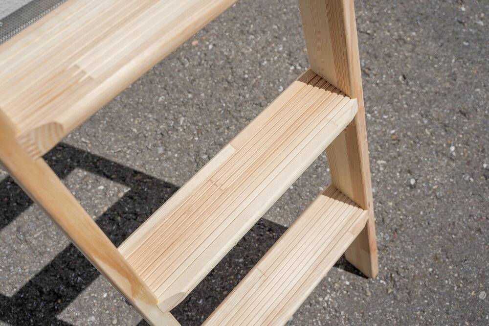 Eine Holzleiter mit hellbraunen Stufen steht auf einem Asphaltuntergrund. Sie hat drei Stufen, die in der Sonne leuchten.