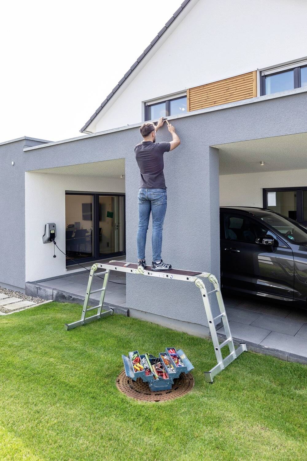 Ein Mann steht auf einer Leiter und repariert das Fenster eines modernen Hauses. Auf dem Rasen befindet sich eine geöffnete Werkzeugkiste.
