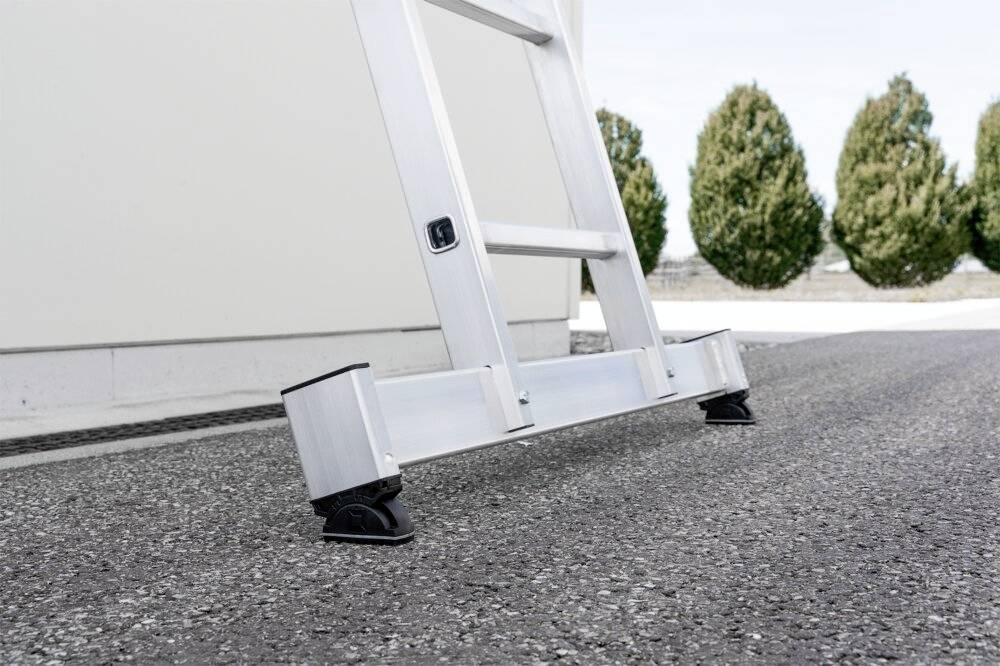 Eine Aluminiumleiter steht auf einem Asphaltboden, gestützt von stabilen Gummifüßen. Im Hintergrund sind Bäume erkennbar.