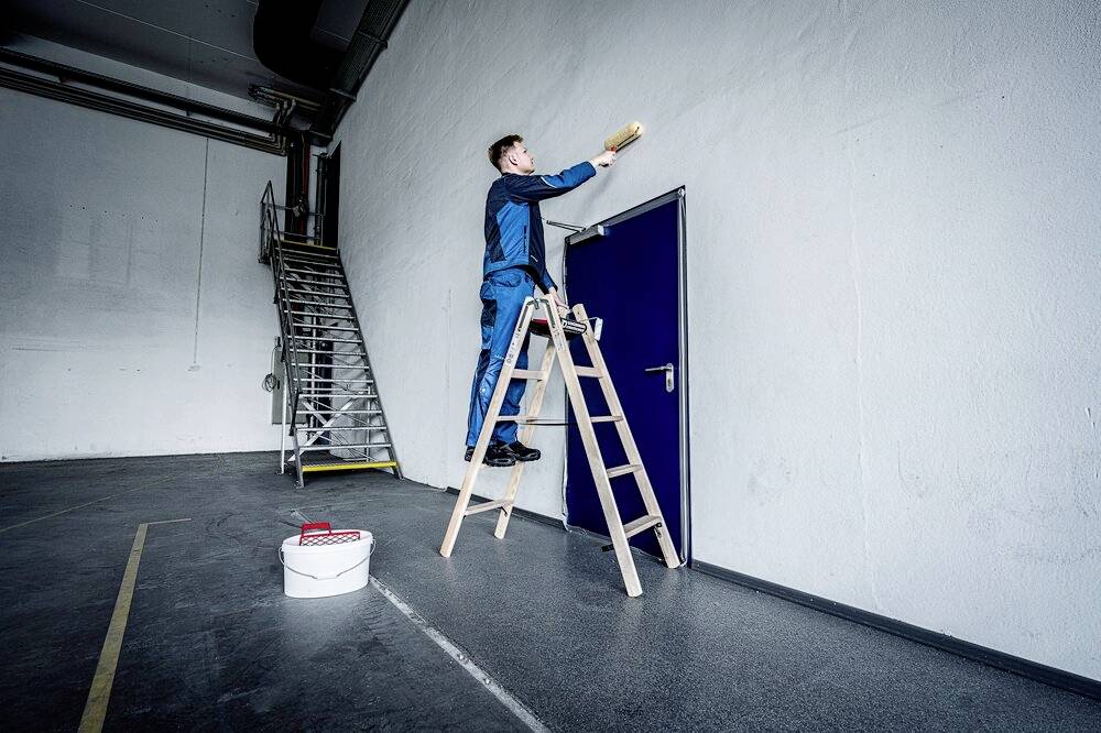 Ein Mann auf einer Leiter streicht eine Wand in einem großen Raum. Eine Farbdose steht am Boden neben einer weiteren Leiter.