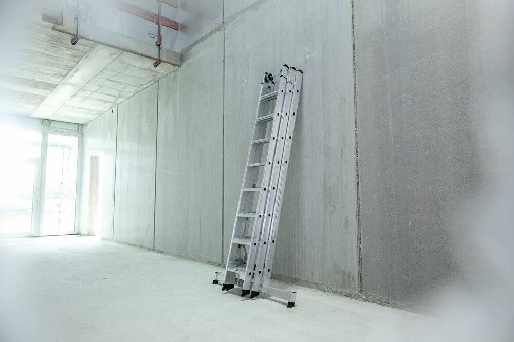 Aluminiumleiter lehnt an einer Betonwand in einem leeren Raum mit Betonboden. Helles Licht strömt durch Glasöffnung links.