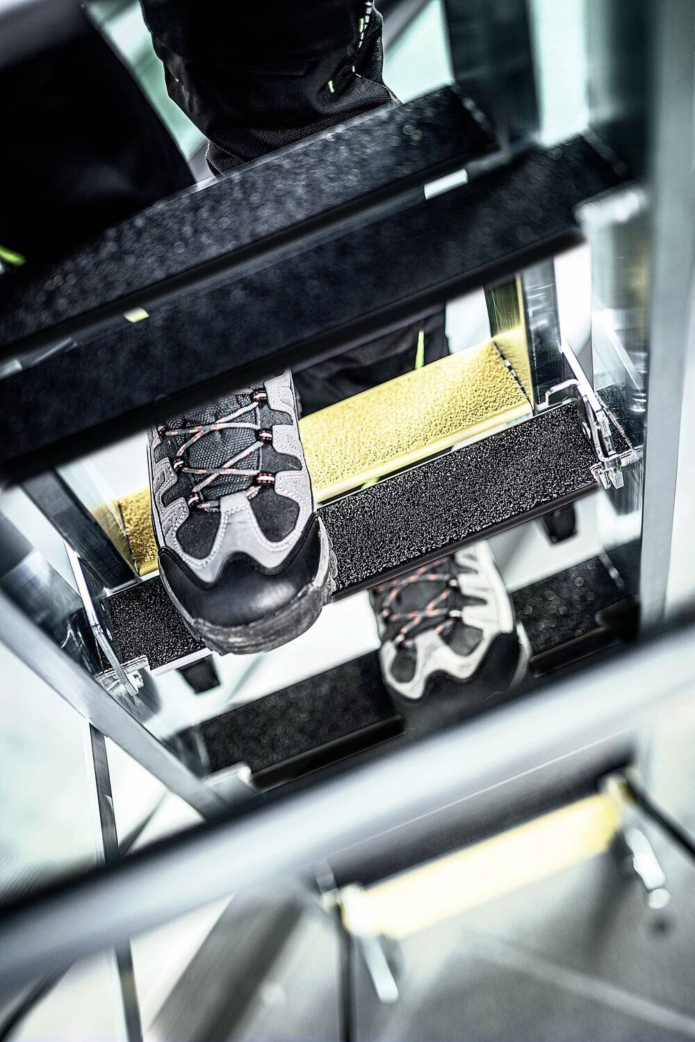 Person in Sicherheitsschuhen steigt eine Metallleiter hinauf. Die Leiter hat gelbe Rutschschutzstreifen auf den Stufen.