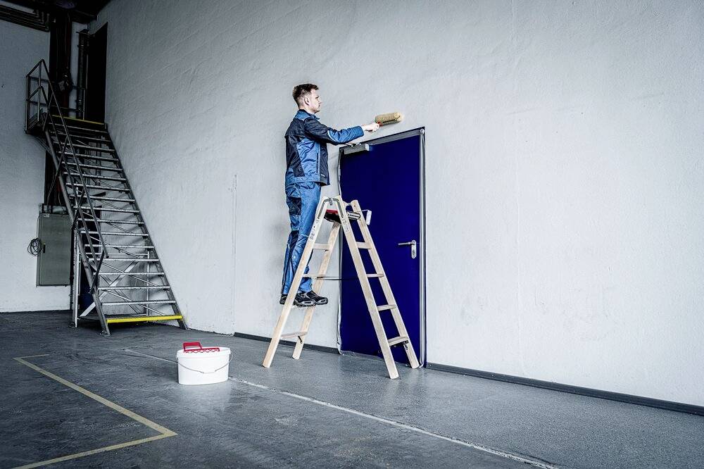 Ein Mann in blauer Arbeitskleidung steht auf einer Leiter und streicht die Wand um eine blaue Tür. Ein Farbeimer steht auf dem Boden.