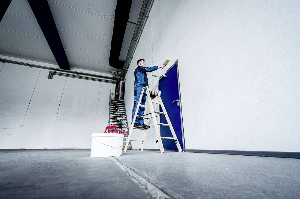 Eine Person steht auf einer Leiter und streicht eine große weiße Wand blau. Im Vordergrund steht ein Farbeimer.