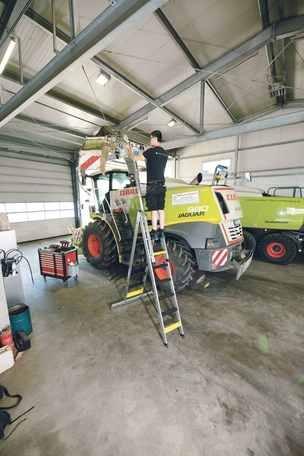 Ein Mechaniker steht auf einer Leiter und arbeitet an einem großen Traktor in einer Werkstatt, umgeben von Werkzeugen und Geräten.