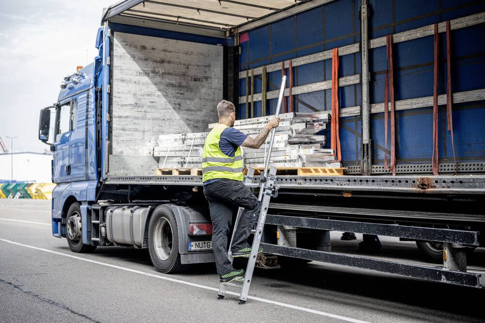 Eine Person in Sicherheitsweste klettert auf eine Leiter an einem geöffneten Lkw, um Metallstangen im Laderaum zu organisieren.
