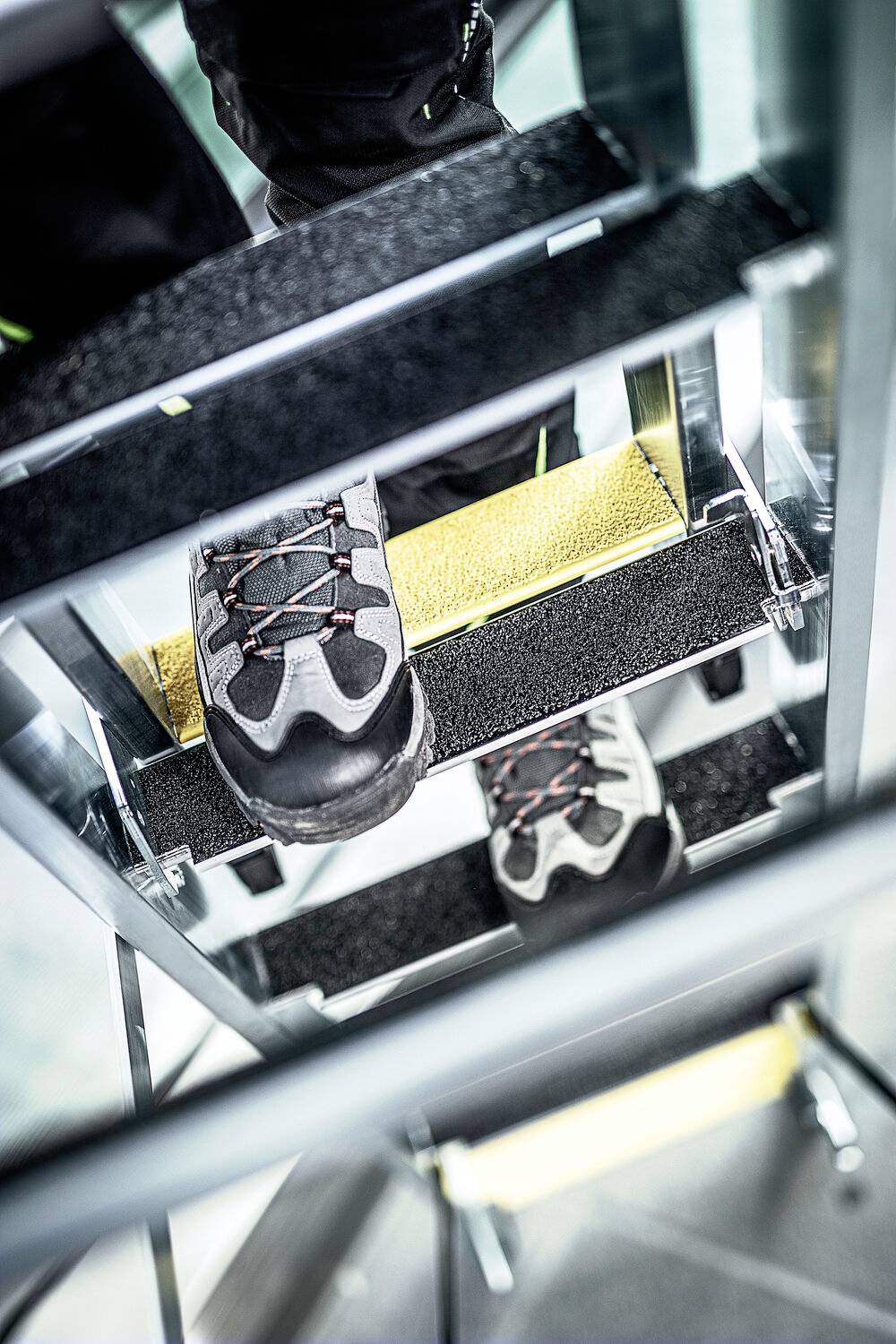 Blick von oben auf eine Person mit Sicherheitsschuhen, die auf einer Metalltreppe steht. Gelbe Anti-Rutsch-Streifen auf den Stufen.