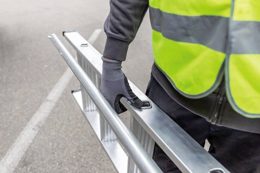 Eine Person in Arbeitskleidung und Sicherheitsweste trägt eine Metallleiter auf einer Straße. Sie trägt Handschuhe für den Schutz.